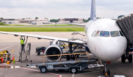 A man in a neon vest stands under the wing of a commercial plane to fill its fuel tank.