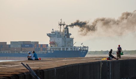 Smoke pours from a cargo ship sailing past a group of fishermen on a pier.