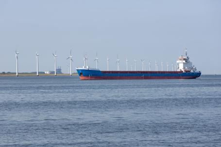 A large cargo ship sails past a row of windmills on shore.