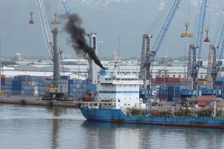 A cargo ship pulls away from a busy shipping port with dark smoke coming from its smokestack.