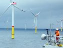 A worker in an orange jumpsuit and white hard hat looks off the side of a ship toward large wind turbines in the ocean.